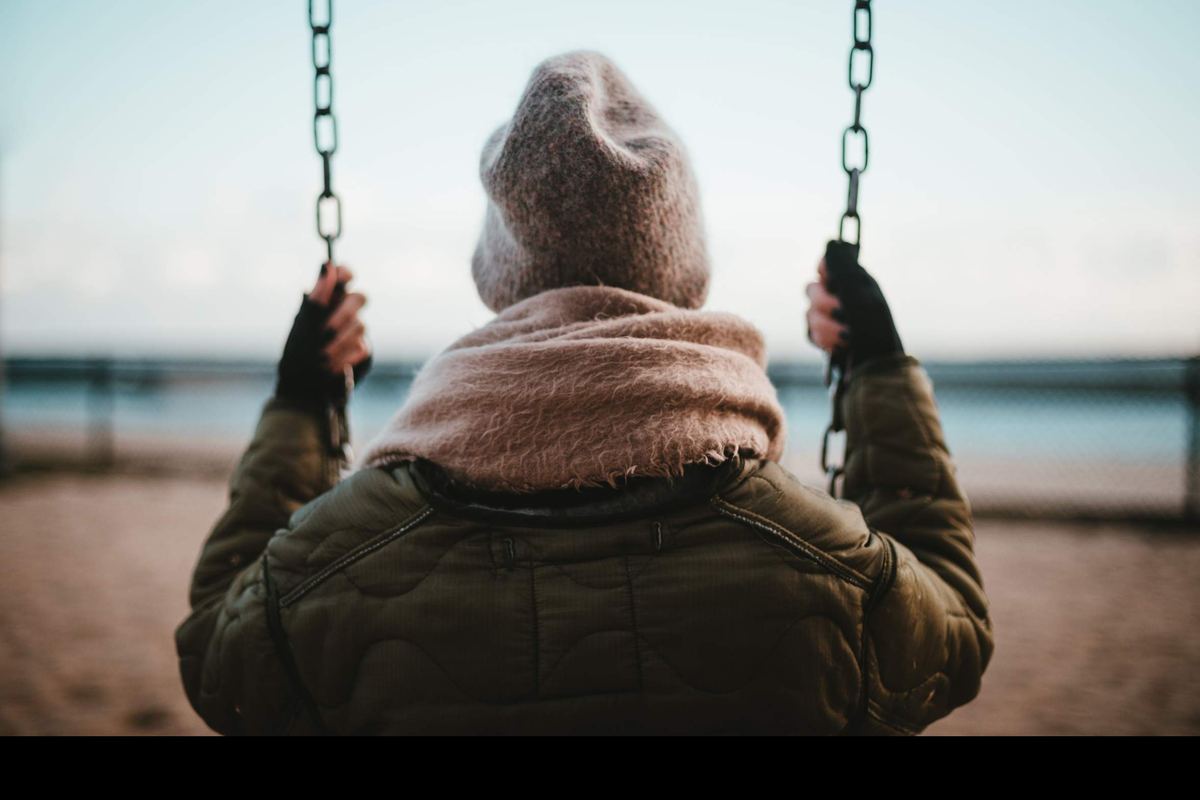 A person in a warm jacket sits alone on a swing and looks out at the sea